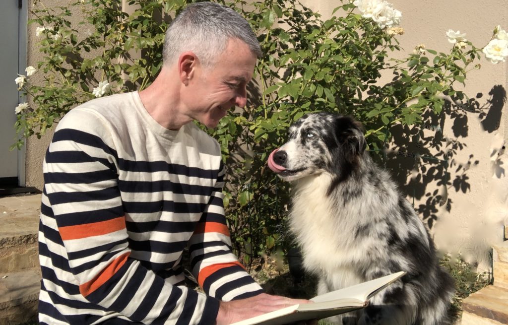 A man in a striped sweater smiles at a dog while reading a book outdoors, surrounded by blooming flowers, capturing a moment of joy and companionship.