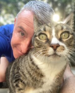 A man smiles close to a tabby cat, both captured in a moment of affection outdoors. Natural light adds warmth to the scene.