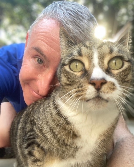 A man smiles close to a tabby cat, both captured in a moment of affection outdoors. Natural light adds warmth to the scene.