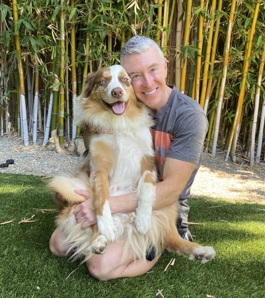 Man smiles while holding a happy, fluffy dog in a lush garden with bamboo in the background. The image captures their joyful bond.