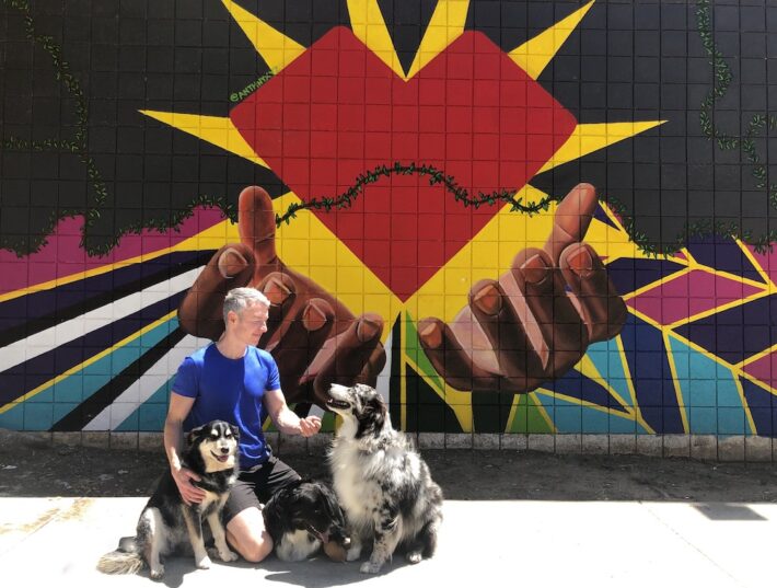 Man kneels with two dogs in front of a vibrant mural featuring hands and a heart, symbolizing love and community.