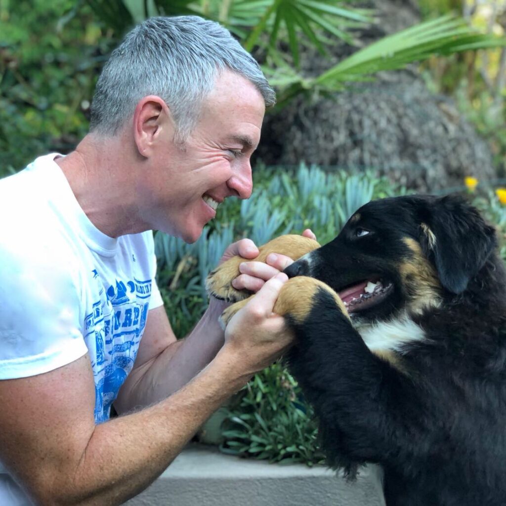 A smiling man plays with a puppy, holding its paws, against a lush green backdrop. Their joyful interaction highlights the bond between humans and pets.