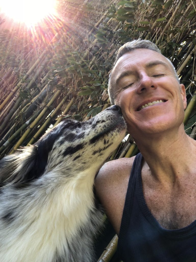 Man smiles blissfully as his dog affectionately licks his cheek in a sunlit bamboo setting, highlighting their bond and joy.