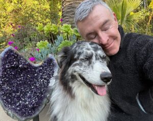 A person with short gray hair embraces a happy, fluffy dog against a backdrop of vibrant plants and a heart-shaped amethyst geode.