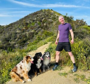 Man in purple shirt and shorts stands on a hiking trail surrounded by four dogs against a scenic hill backdrop. Perfect for outdoor activity content.