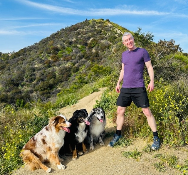 Man in purple shirt and shorts stands on a hiking trail surrounded by four dogs against a scenic hill backdrop. Perfect for outdoor activity content.