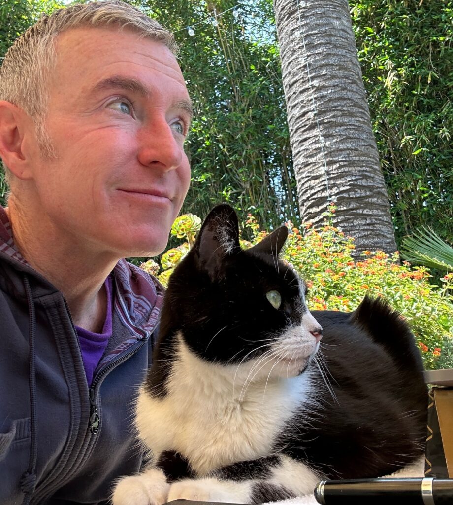 Man and a black-and-white cat sit together outdoors, surrounded by greenery. Their expressions suggest a quiet moment of companionship.