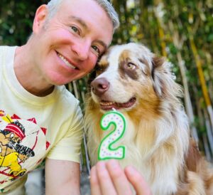 A smiling individual poses with a fluffy dog, holding a green number "2" decoration, celebrating the dog's birthday in a lush outdoor setting.