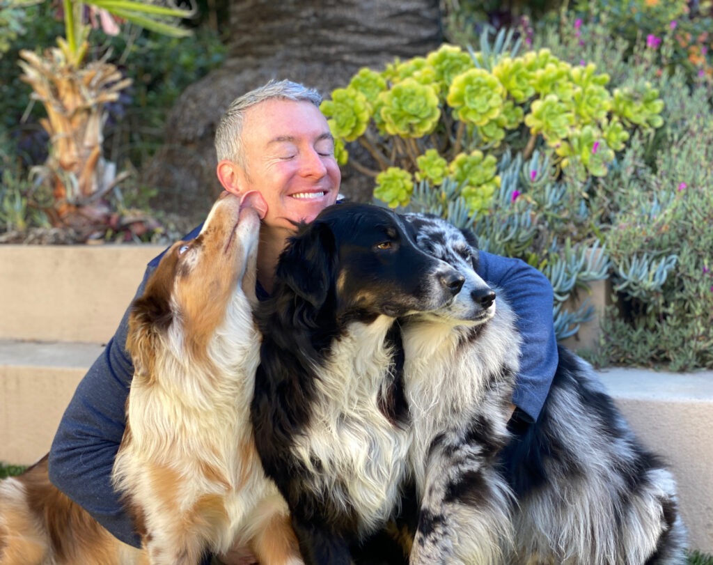 A person smiles with closed eyes, embraced by two dogs, one licking their cheek. Lush greenery in the background enhances the joyful scene.
