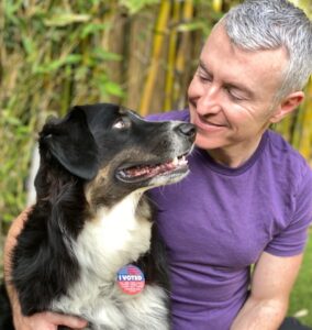 A man wearing a purple shirt smiles at his dog, who proudly displays an "I Voted" sticker. The image highlights the joy of voting companionship.