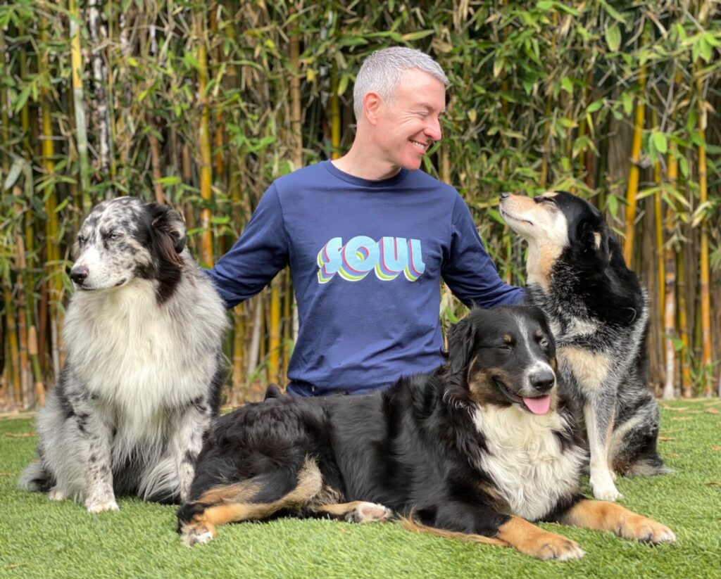 A man smiles while seated on grass with three dogs, showcasing a joyful moment against a lush bamboo backdrop.