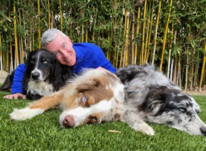 A man in a blue sweater smiles while lying on the grass with three dogs, showcasing their bond in a tranquil, green garden setting.