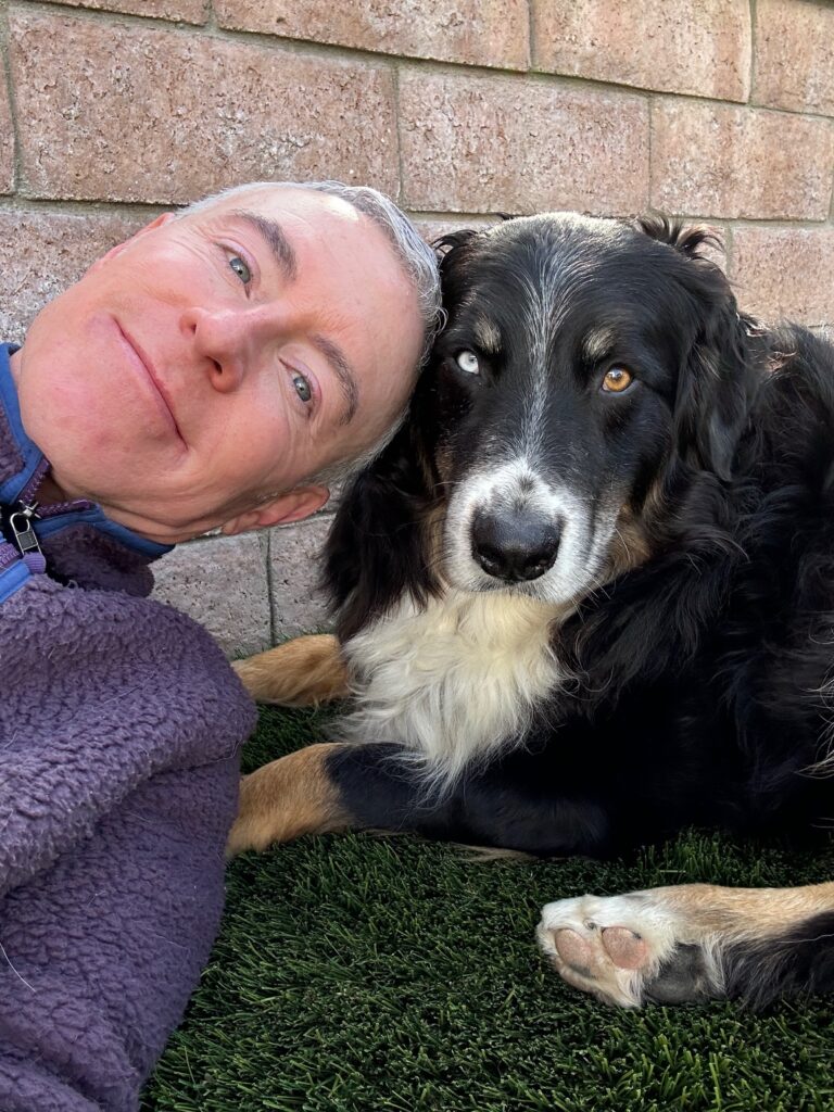 A person smiles while resting next to a black and tan dog on artificial grass, showcasing their bond and a moment of relaxation together.