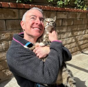 A smiling person cuddles a tabby cat in a sunlit outdoor space, showcasing a warm bond between them in a serene setting.