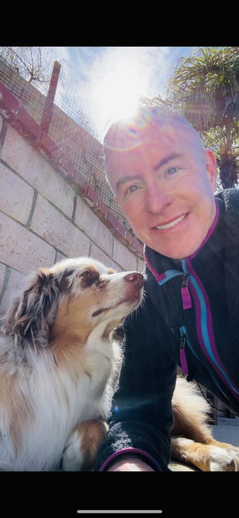 A person smiles while posing with an Australian Shepherd dog in a sunny outdoor setting, showcasing their bond in a relaxed atmosphere.