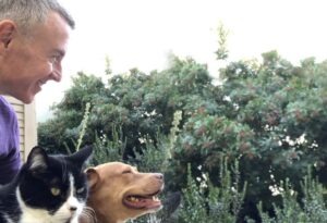 Man smiling at a dog and cat outdoors, surrounded by greenery. The image highlights companionship and a joyful moment with pets.