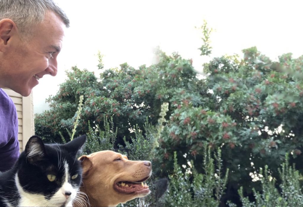 Man smiling at a dog and cat outdoors, surrounded by greenery. The image highlights companionship and a joyful moment with pets.