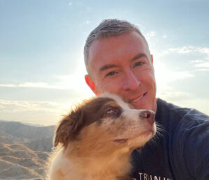 Smiling man with a light gray dog in a selfie against a scenic backdrop of mountains and sky, emphasizing companionship and outdoor adventure.
