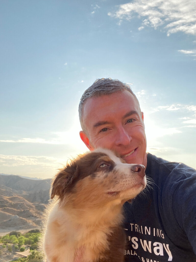 Smiling man with a light gray dog in a selfie against a scenic backdrop of mountains and sky, emphasizing companionship and outdoor adventure.