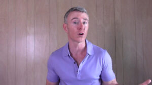 A man in a purple polo shirt gestures while speaking, set against a light wooden panel background. He appears to be presenting or teaching.