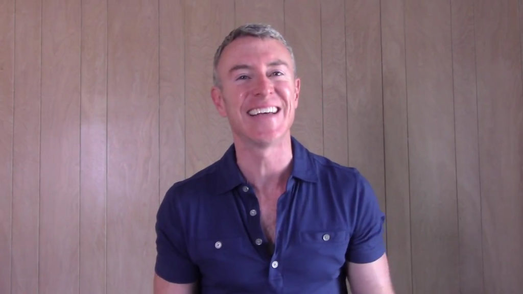Smiling man in a navy blue shirt against a light wooden background, possibly conveying a positive message or personal introduction.