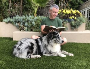 A man reads a book while sitting on grass next to a large, fluffy, black-and-white dog. Lush greenery and succulents frame the cozy outdoor scene.