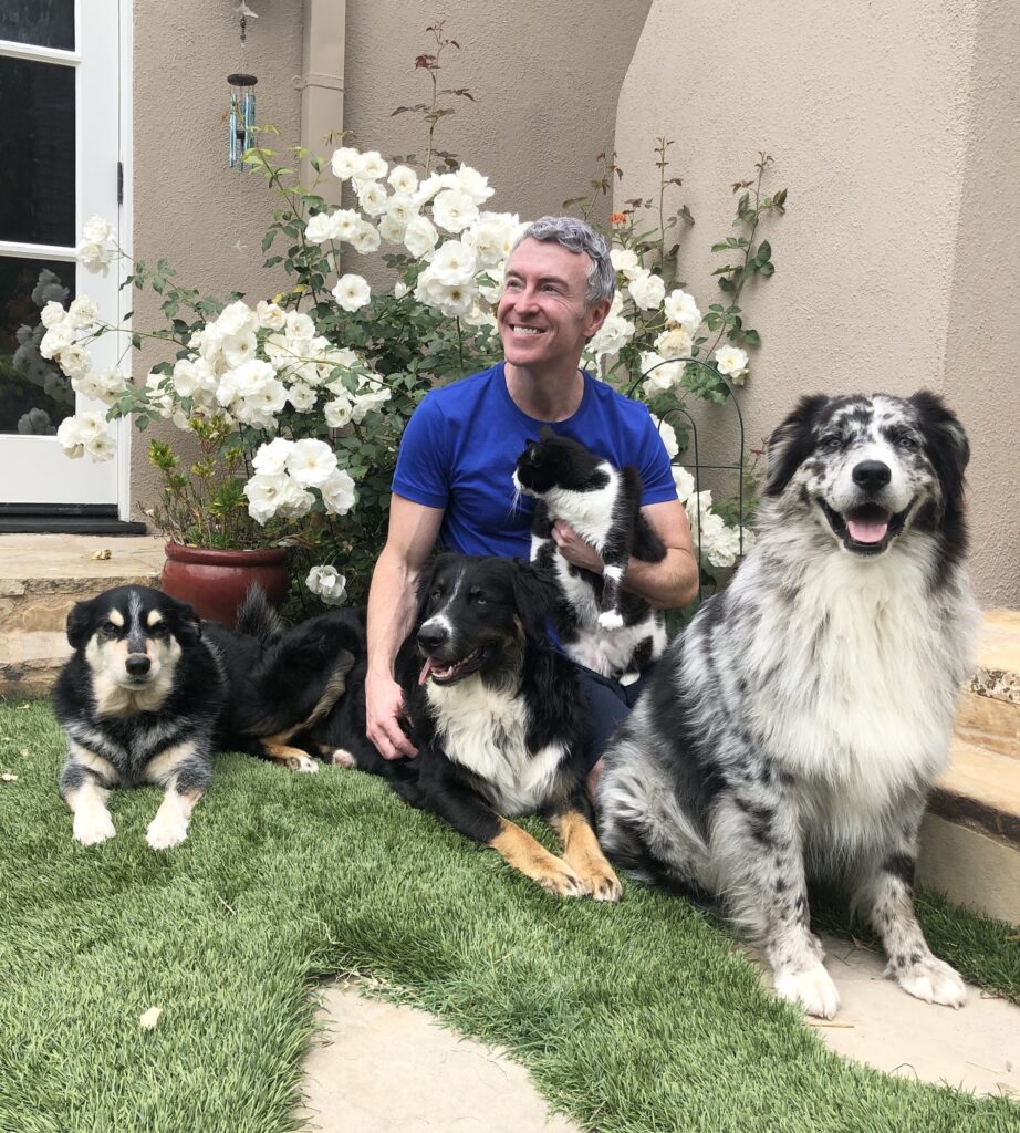 A smiling man sits among four pets—a black dog, a fluffy blue merle dog, and two cats—surrounded by white roses and a cozy home setting.