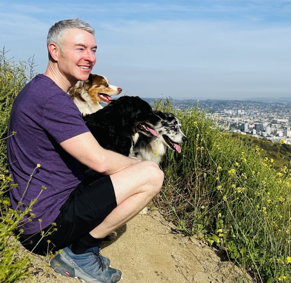 A man squats on a hillside, smiling with three dogs beside him, overlooking a cityscape below. The scene captures a moment of joy in nature.