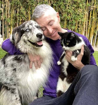 Man joyfully embraces a large, fluffy dog and a black-and-white cat in a lush, green outdoor setting, surrounded by bamboo.